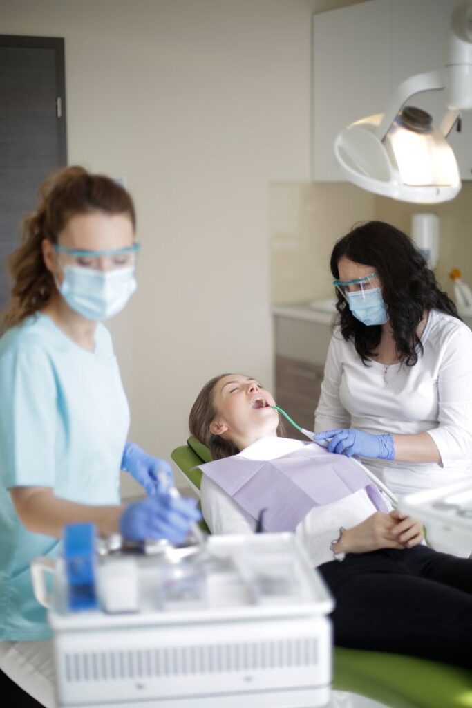 Young female dentist in mask latex gloves and protective glasses wearing uniform with assistant using modern equipment and dental tools while curing teeth of female patient in dental clinic